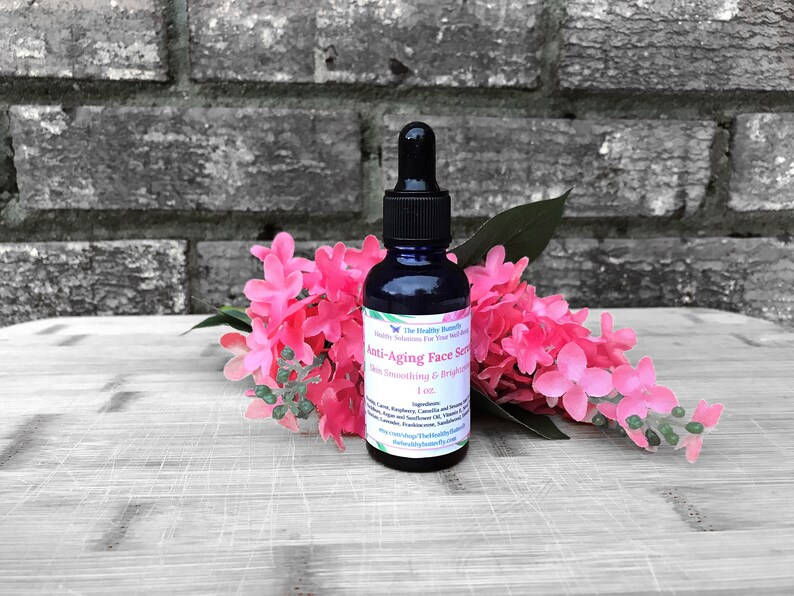 May include: A dark blue glass bottle of anti-aging face serum with a black dropper, set against a backdrop of pink flowers and a gray brick wall. The label reads "Anti-Aging Face Serum, Skin Smoothing & Brightening."