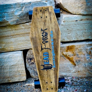 Könnte beinhalten: Ein Longboard aus Holz mit schwarzen Trucks und Rädern. Das Deck ist braun gebeizt und hat ein schwarz-weißes Grafikmotiv eines Käfers mit dem Text "DEAD FOREST longboards".