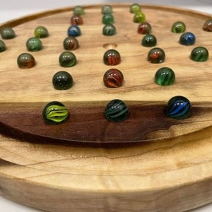 Peut inclure: Un jeu de solitaire en bois avec une finition marron clair. Le plateau comporte une série de trous remplis de billes colorées dans les tons de vert, rouge, bleu et noir. Les billes sont disposées en cercle.