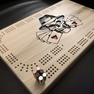 May include: A wooden cribbage board with a skull and playing cards design. The board is light-colored with a dark engraved skull wearing a hat and holding cards with red hearts. Six metal pegs are in the foreground.