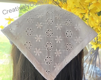 May include: White eyelet headscarf with a floral snowflake pattern. The square scarf is worn on the head, with the text "Creatively Damaris" visible in the upper left corner. Yellow flowers are in the background.
