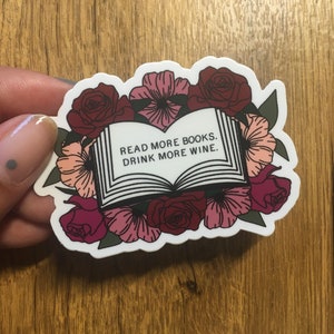 May include: A sticker featuring an open book illustration surrounded by burgundy and pink flowers. The text on the book reads, "READ MORE BOOKS. DRINK MORE WINE."