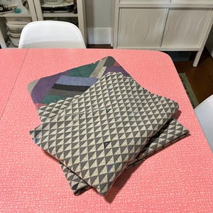 May include: Two folded fabric napkins with geometric patterns rest on a pink table. One napkin features a gray and white triangle design, while the other has a colorful, abstract pattern with purple, green, and blue hues. The table has a textured surface.