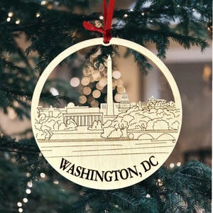 Puede incluir: Un adorno navideño redondo de madera con un diseño cortado con láser del horizonte de Washington, D.C., que incluye el Monumento a Washington y el Capitolio. El adorno está suspendido por una cinta roja y tiene grabadas las palabras "WASHINGTON, DC".