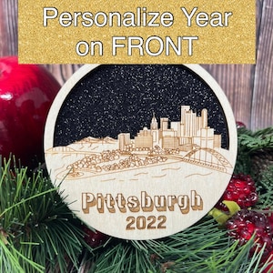 May include: A wooden bauble featuring a black and white silhouette of the Pittsburgh skyline with the text "Pittsburgh 2022". The bauble is surrounded by red apples and evergreen branches.