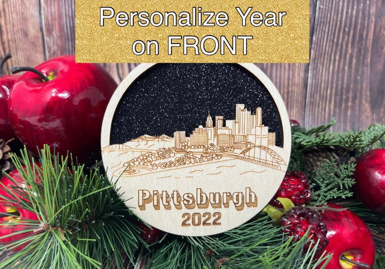 May include: A wooden bauble featuring a black and white silhouette of the Pittsburgh skyline with the text "Pittsburgh 2022". The bauble is surrounded by red apples and evergreen branches.
