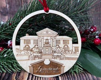 May include: A wooden Christmas ornament shaped like a circle with a white outline of Graceland, Elvis Presley's home, in the center. The ornament has the text "Graceland, Home of Elvis A. Presley, 37627" at the bottom.