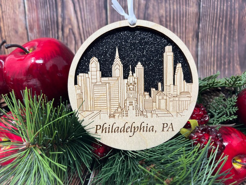 May include: A round wooden bauble with a black glitter background and a white silhouette of the Philadelphia skyline. The text "Philadelphia, PA" is written below the skyline.