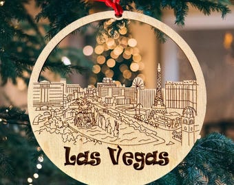 May include: A wooden Christmas ornament featuring a laser-cut design of the Las Vegas skyline. The ornament is round and has a red ribbon attached to the top. The text "Las Vegas" is printed on the bottom of the ornament.