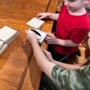 Puede incluir: Dos ni&ntilde;os est&aacute;n sentados en una mesa de madera, uno sostiene una peque&ntilde;a caja con un adorno blanco y negro en su interior. Hay varias otras cajas apiladas en la mesa.