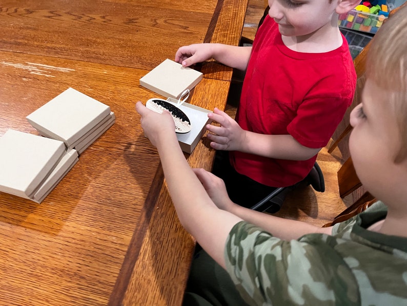 Puede incluir: Dos ni&ntilde;os est&aacute;n sentados en una mesa de madera, uno sostiene una peque&ntilde;a caja con un adorno blanco y negro en su interior. Hay varias otras cajas apiladas en la mesa.