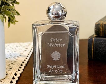 Puede incluir: Una botella de cristal transparente con un tapón esférico plateado. El frente está grabado con el texto Peter Webster, un icono de concha y el texto Baptized 8/17/25. La botella tiene un cuerpo rectangular con bordes biselados.