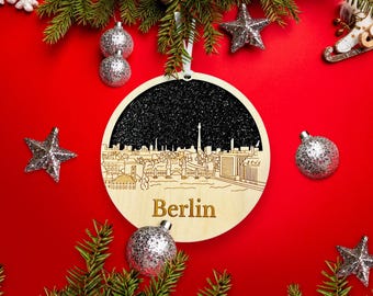 Könnte beinhalten: Weihnachtsschmuck aus Holz mit einem lasergeschnittenen Skyline von Berlin, Deutschland, und dem Namen der Stadt in goldfarbenen Buchstaben. Der Schmuck hat einen schwarzen Glitzerhintergrund, umgeben von silbernen Kugeln und Sternen auf rotem Hintergrund.