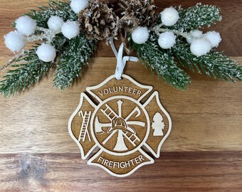 May include: A wooden Christmas ornament shaped like a firefighter's Maltese cross. The ornament is painted brown and features a white outline of the cross, a firefighter's helmet, an axe, a ladder, and a fire hydrant. The words "Volunteer Firefighter" are written on the ornament.