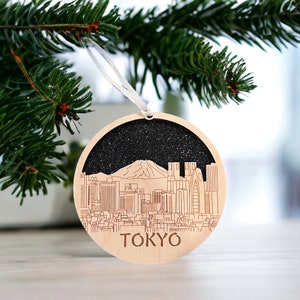 Puede incluir: Un adorno navide&ntilde;o de madera con una silueta negra del horizonte de Tokio con el monte Fuji en el fondo. El adorno est&aacute; decorado con purpurina y la palabra "TOKIO" est&aacute; inscrita debajo del horizonte.