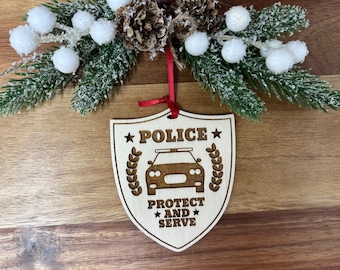Könnte beinhalten: Weihnachtsbaumschmuck aus Holz in Form eines Schildes mit den Worten "Police Protect and Serve" und einer Polizeiautosilhouette. Der Schmuck ist mit Sternen und einem Kranz verziert.