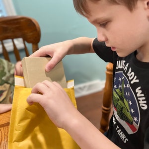 Puede incluir: Un ni&ntilde;o est&aacute; abriendo un sobre amarillo con un libro marr&oacute;n dentro. El ni&ntilde;o lleva una camiseta negra con el texto "Just a boy who loves tanks" y un gr&aacute;fico de un tanque verde.