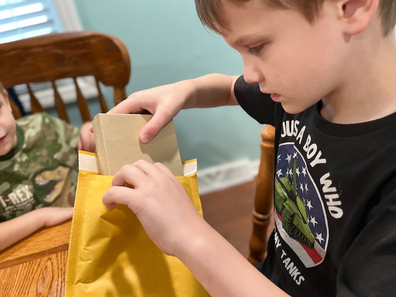 Puede incluir: Un ni&ntilde;o est&aacute; abriendo un sobre amarillo con un libro marr&oacute;n dentro. El ni&ntilde;o lleva una camiseta negra con el texto "Just a boy who loves tanks" y un gr&aacute;fico de un tanque verde.