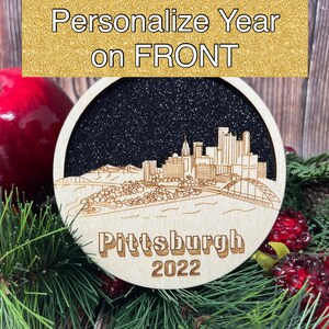 Puede incluir: Un adorno navide&ntilde;o de madera con una silueta en blanco y negro del horizonte de Pittsburgh con el texto "Pittsburgh 2022". El adorno tiene un fondo negro brillante.