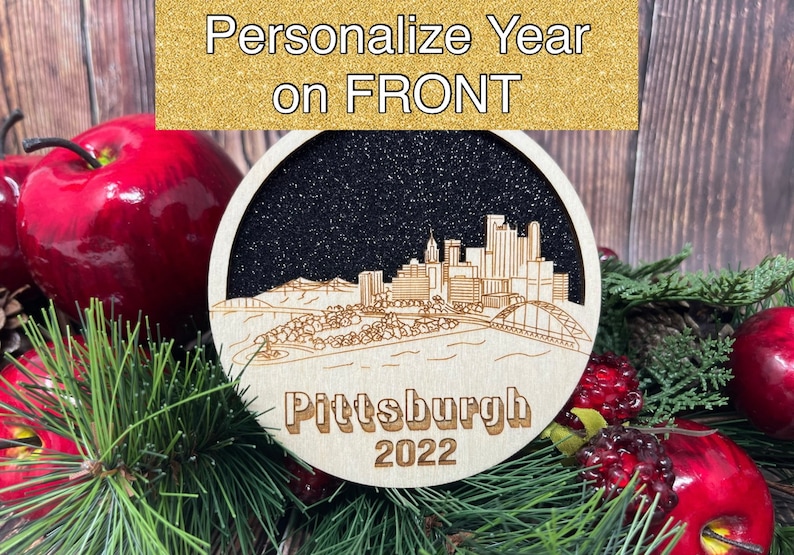 Puede incluir: Un adorno navide&ntilde;o de madera con una silueta en blanco y negro del horizonte de Pittsburgh con el texto "Pittsburgh 2022". El adorno tiene un fondo negro brillante.