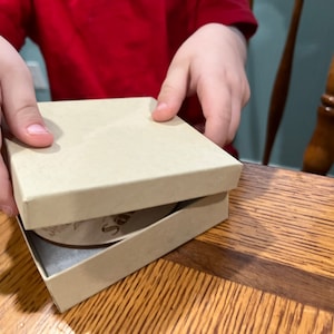 Puede incluir: Las manos de un ni&ntilde;o abren una peque&ntilde;a caja beige que revela un adorno redondo de madera con la palabra "Santa" grabada.