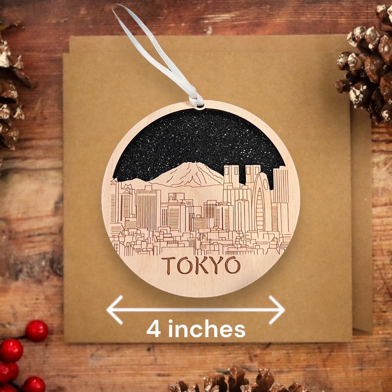 Puede incluir: Un adorno redondo de madera con fondo de purpurina negra y un contorno blanco del horizonte de Tokio, incluyendo el monte Fuji. El adorno tiene un di&aacute;metro de 10 cm y la palabra "TOKIO" escrita debajo del horizonte.
