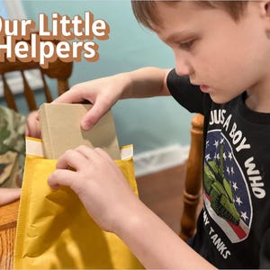 May include: A young person is carefully removing a rectangular, beige package from a yellow padded envelope. The text "Our Little Helpers" is displayed in a playful font at the top. The person is wearing a black t-shirt with a military-themed graphic.