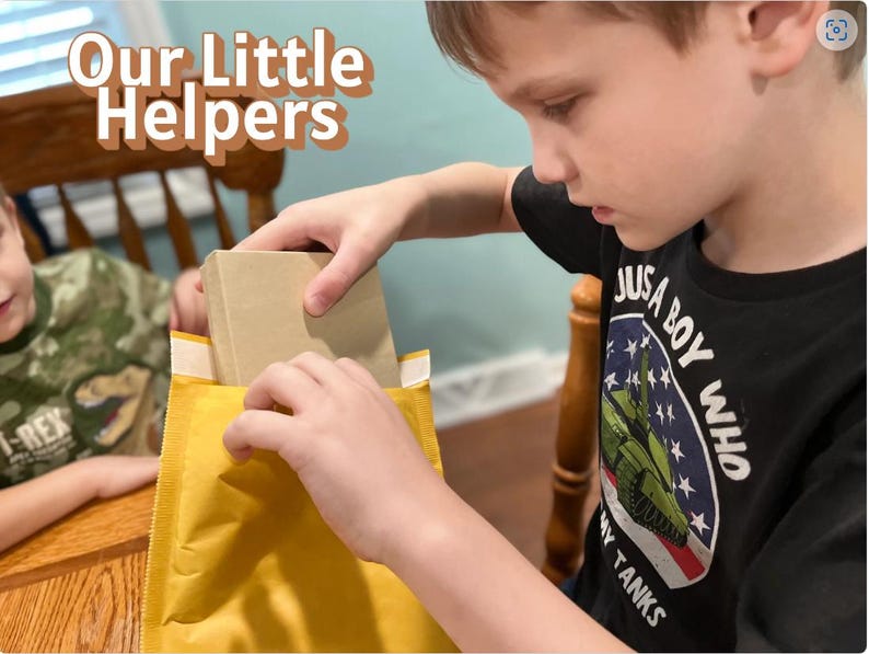 May include: A young person is carefully removing a rectangular, beige package from a yellow padded envelope. The text "Our Little Helpers" is displayed in a playful font at the top. The person is wearing a black t-shirt with a military-themed graphic.