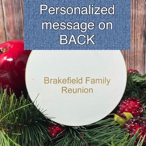 May include: A white round wooden sign with the text "Brakefield Family Reunion" in gold lettering. The sign is surrounded by red apples, pine branches, and red berries.