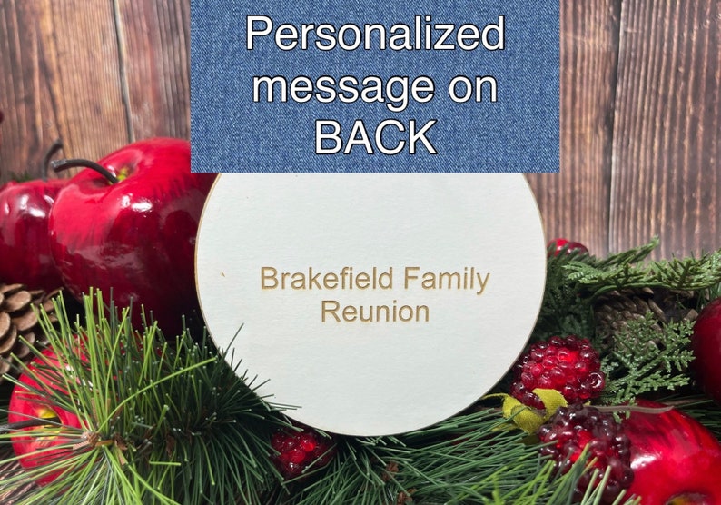 May include: A white round wooden sign with the text "Brakefield Family Reunion" in gold lettering. The sign is surrounded by red apples, pine branches, and red berries.