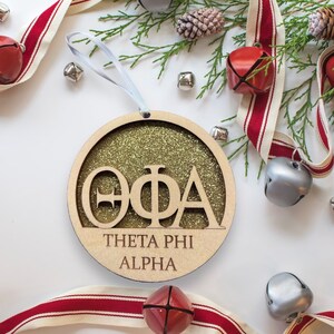 May include: A round, wooden ornament with gold glitter. The ornament features the Greek letters "ΘΦΑ" in a cutout design, with the words "Theta Phi Alpha" written below.