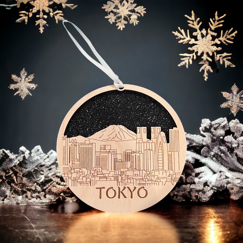 Puede incluir: Un adorno navide&ntilde;o de madera redondo con un fondo de purpurina negra y una silueta del horizonte de Tokio, incluyendo el monte Fuji. El adorno tiene una cinta blanca y el texto "TOKYO" est&aacute; impreso en la parte inferior.
