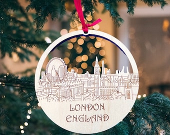 May include: A wooden Christmas ornament featuring a detailed silhouette of the London skyline, including the London Eye, Big Ben, and other iconic landmarks. The ornament is painted in white and has the text "LONDON ENGLAND" printed on the bottom.