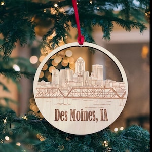 以下が含まれることがあります： Des Moines, IAの街並みを細部まで描いたシルエットが刻まれた、丸い木製クリスマスオーナメント。オーナメントには赤いリボンが付いています。