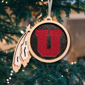 May include: A wooden Christmas ornament featuring a red "U" with black glitter on a black background. The ornament is shaped like a feather and has a white string for hanging.