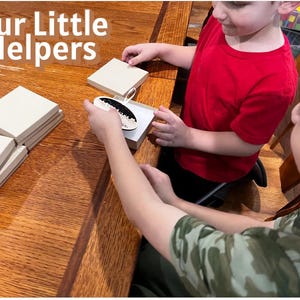 May include: Two children are working with small, square, light-colored boxes and a decorative item. The text "Our Little Helpers" is displayed in white letters at the top left of the image. The scene takes place on a wooden table.