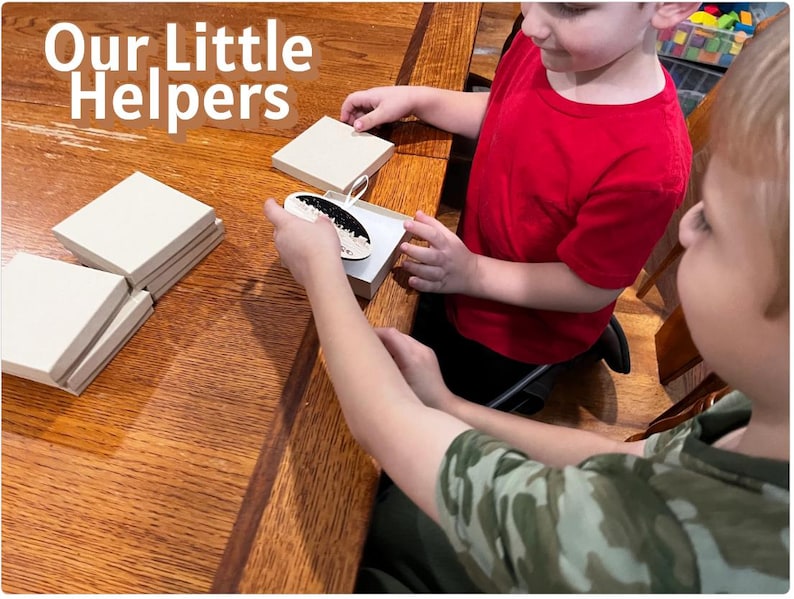 May include: Two children are working with small, square, light-colored boxes and a decorative item. The text "Our Little Helpers" is displayed in white letters at the top left of the image. The scene takes place on a wooden table.