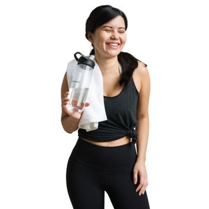 May include: A woman wearing black workout clothes is holding a clear water bottle with a black lid and a white towel draped over her shoulder. She is smiling.