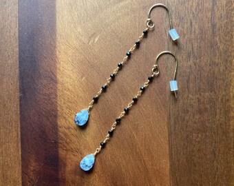 May include: A pair of gold-colored earrings with a delicate design. Each earring features a chain of small black beads and a teardrop-shaped, light blue stone at the bottom. The earrings are displayed on a wooden surface.
