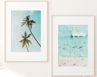 May include: Two framed prints, one with a beach scene and the other with palm trees against a blue sky. The prints are hanging on a white wall above a wooden cabinet with a wicker basket and a potted plant.
