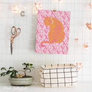 Op de afbeelding: Een roze en oranje tijgerprint met een roze achtergrond met een herhalend patroon van harten en de letters XO. De print hangt aan een witte rasterwand.