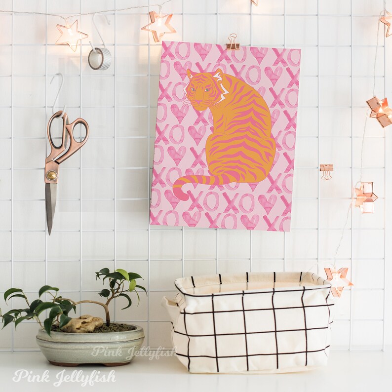 Op de afbeelding: Een roze en oranje tijgerprint met een roze achtergrond met een herhalend patroon van harten en de letters XO. De print hangt aan een witte rasterwand.