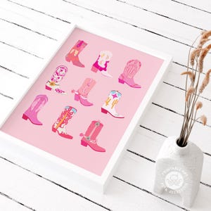 May include: A framed print featuring a collection of colorful cowboy boots in various shades of pink, red, and white. The boots are arranged on a pink background, creating a vibrant and stylish design. The print is in a white frame.