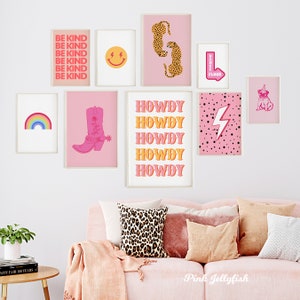 Pode incluir: Uma sala de estar rosa com um sof&aacute; rosa e uma parede de galeria com estampas rosa e amarelas. As estampas incluem um arco-&iacute;ris, uma bota de cowboy, um rosto sorridente, um raio, um leopardo e a palavra "Howdy" repetida cinco vezes.
