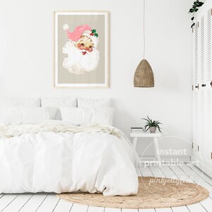 May include: A printable art print featuring a vintage illustration of Father Christmas with a pink hat and a white beard. The print is framed and hanging on a white wall above a white bed with a white duvet and pillows.