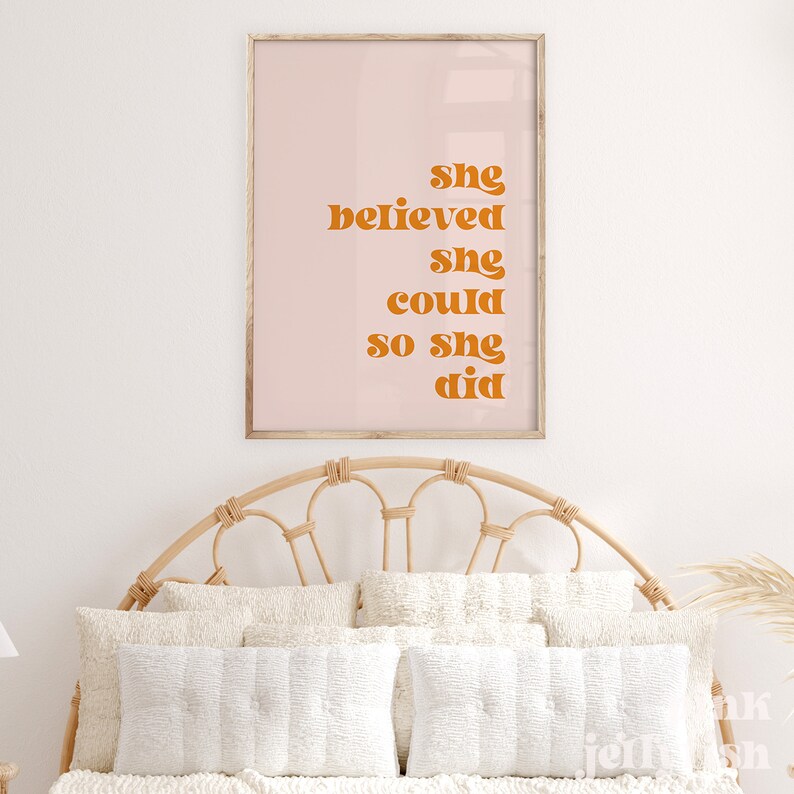 Op de afbeelding: Een ingelijste print met de tekst "She believed she could so she did" in oranje letters op een lichtroze achtergrond. De print wordt weergegeven boven een bed met witte kussens en een geweven hoofdbord.