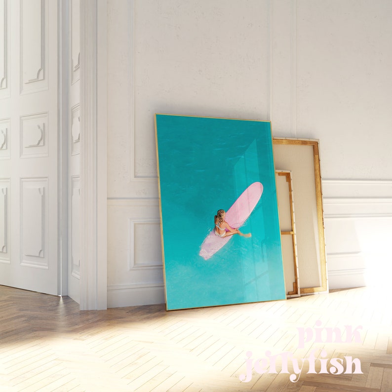 May include: A framed print of a woman on a pink surfboard in turquoise water. The print has a gold frame.