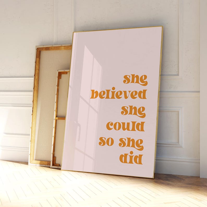 Op de afbeelding: Een ingelijste print met de tekst "She believed she could so she did" in oranje letters. De print leunt tegen een muur met twee andere doeken. De achtergrond is lichtroze.