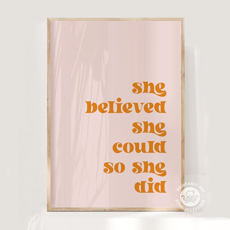 Op de afbeelding: Een ingelijste print met de tekst "She believed she could so she did" in oranje letters op een lichtroze achtergrond. De print heeft een houten lijst.