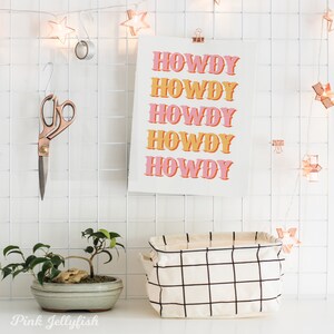 May include: A white grid wall with a pink and orange print that says "Howdy" five times. There is a small potted bonsai tree, a white and black grid storage basket, and a pair of scissors hanging on the wall.